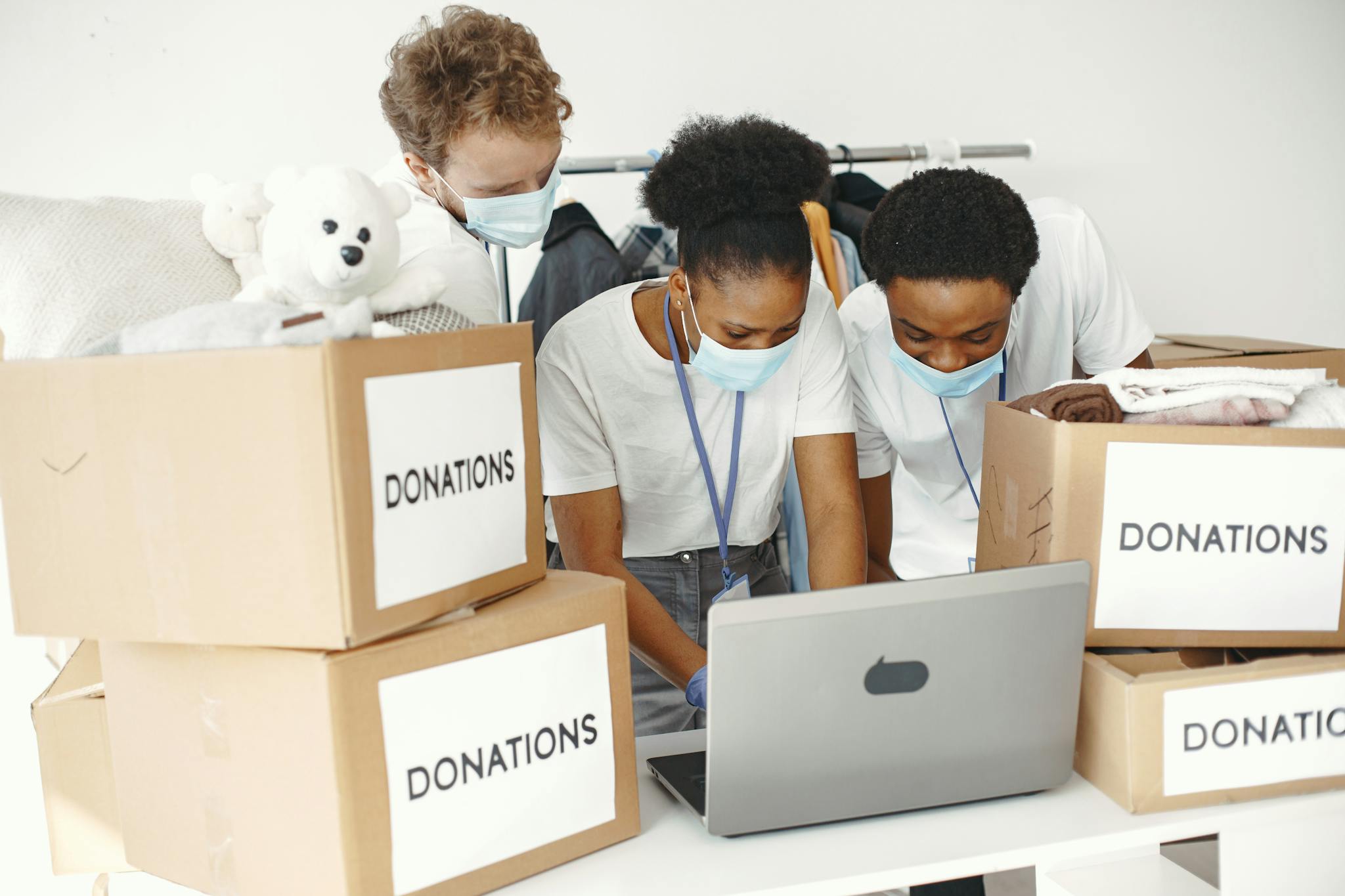 Group of volunteers wearing masks sorting donations with a laptop, promoting diversity and teamwork.